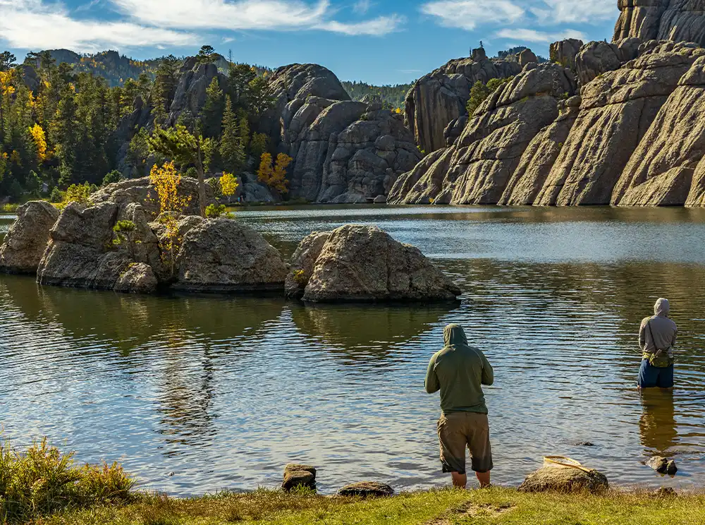 Two people fish by a rocky lake with rugged cliffs and trees under a blue sky.