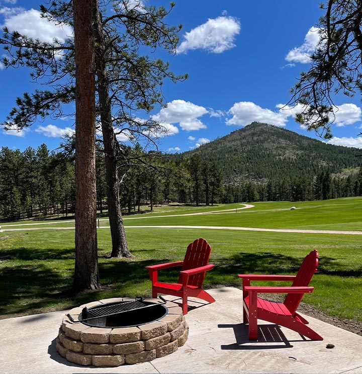 Two red chairs by a fire pit, overlooking a grassy field and pine trees, with a mountain in view.