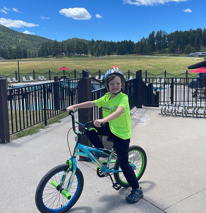 Child in a bright shirt on a bike, near a pool and grassy field with trees in the background.