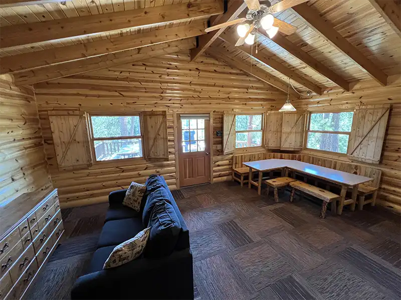 Log cabin interior with a black sofa, wooden table, benches, and a ceiling fan.