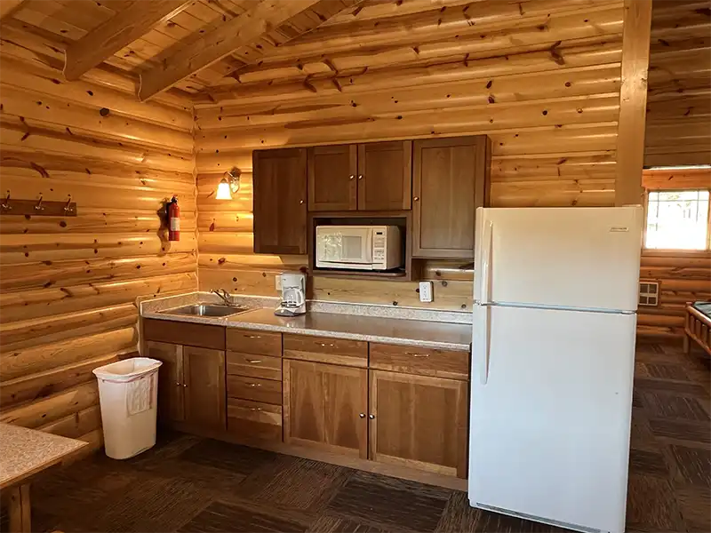 Wooden cabin kitchen with cabinets, microwave, fridge, sink, coffee maker, and trash bin.