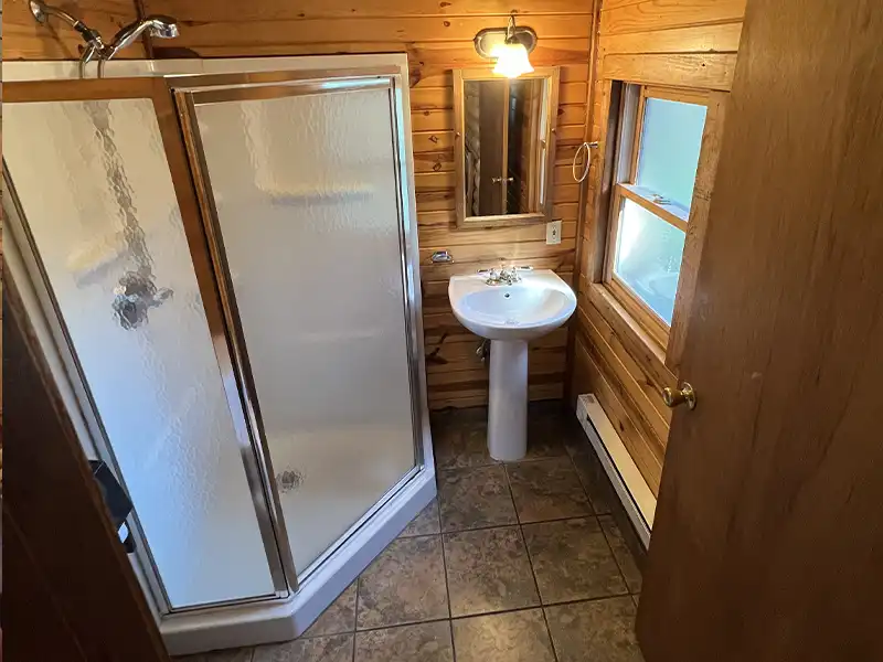 Small wooden bathroom with a shower, sink, mirror, and window.