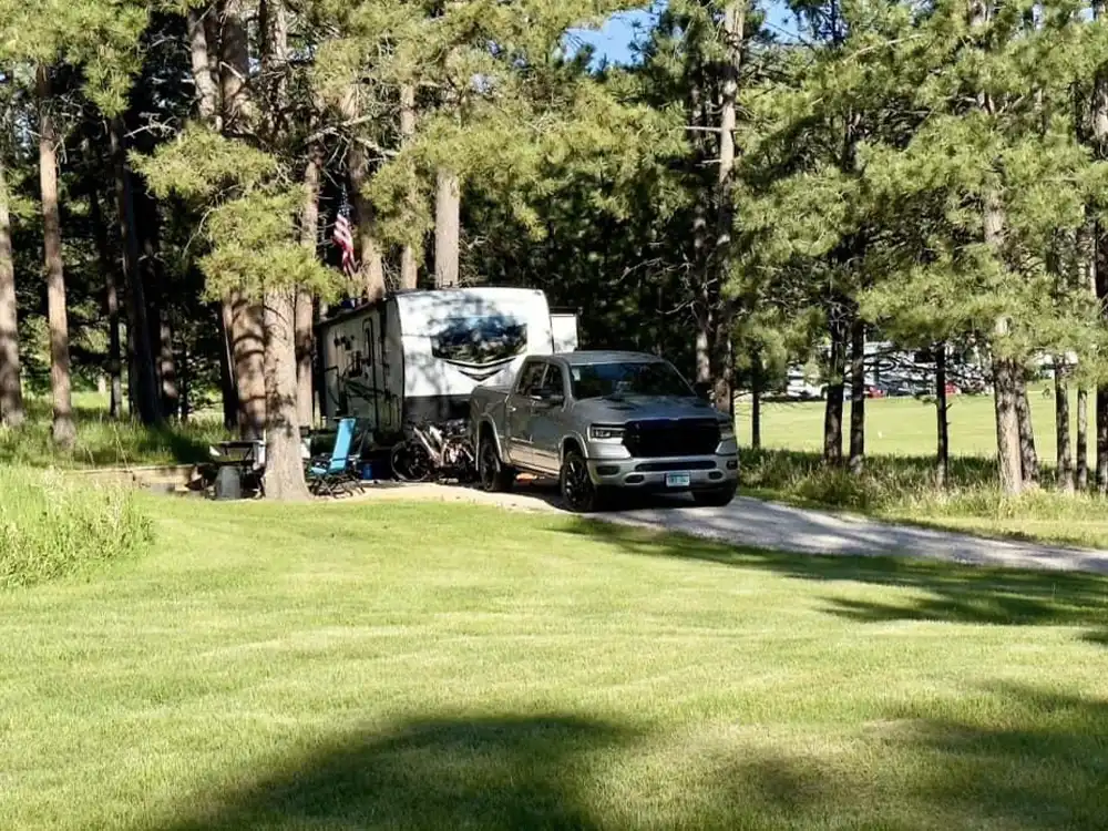 Truck and RV parked in a forested area with lawn chairs nearby.