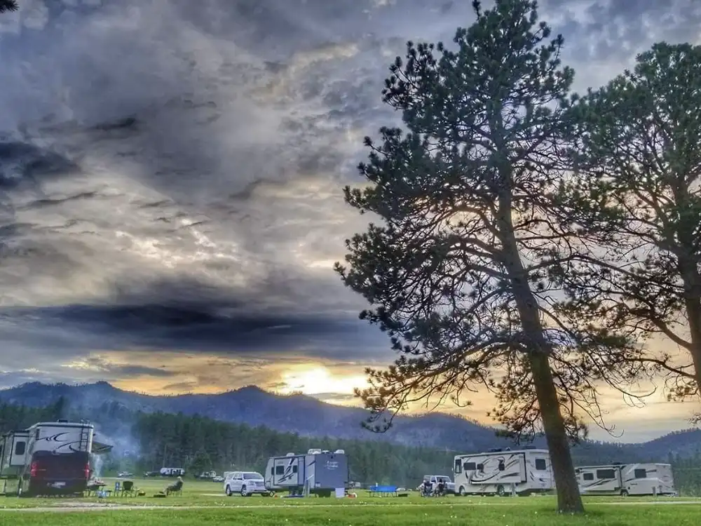 Camping area with RVs, trees, and a dramatic cloudy sky at sunset.