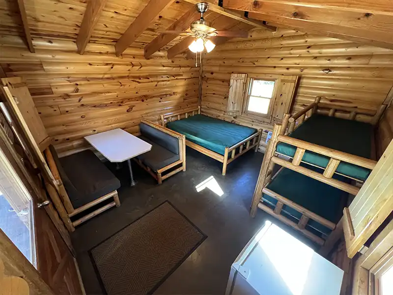 Log cabin interior with wooden furniture, bunk bed, single bed, table, and ceiling fan.