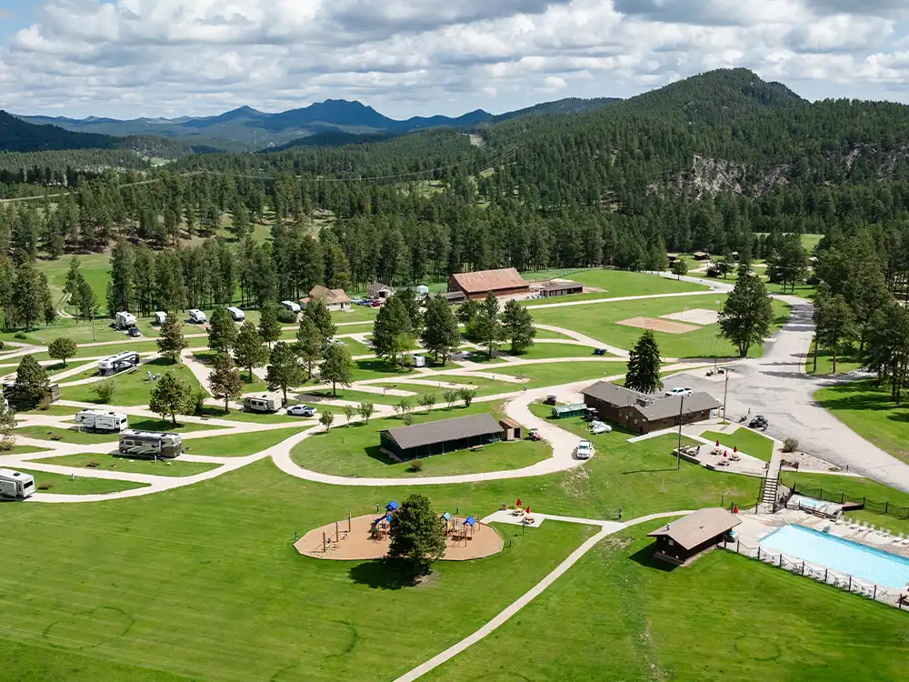 Aerial view of a campground with RVs, a pool, playground, and surrounding forested hills.