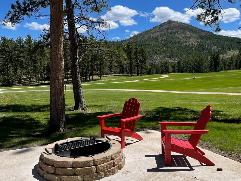 Two red chairs by a fire pit overlook a grassy field and mountain under a blue sky.