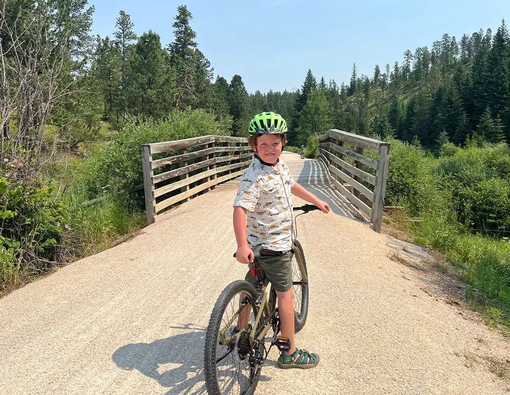 Child with a green helmet on a bike, standing on a bridge in a forest area.