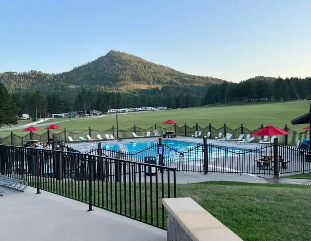 Outdoor pool with red umbrellas, fenced area, grassy field, and mountain in the background.