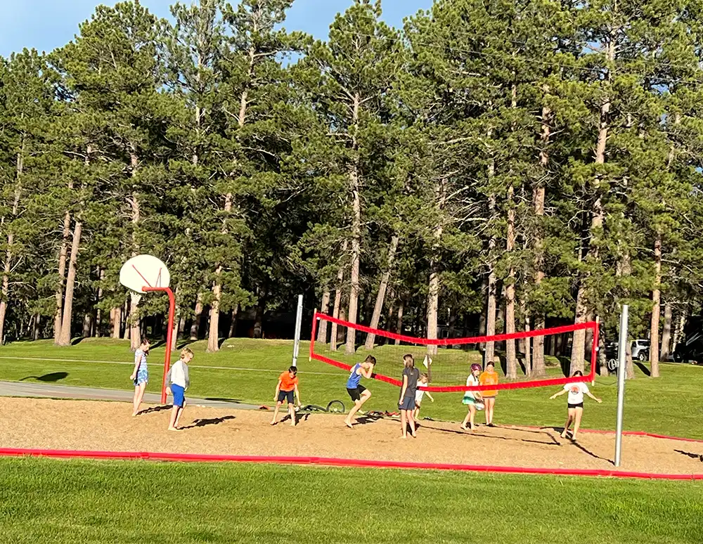 Children play volleyball on a sandy court in a park with trees in the background.