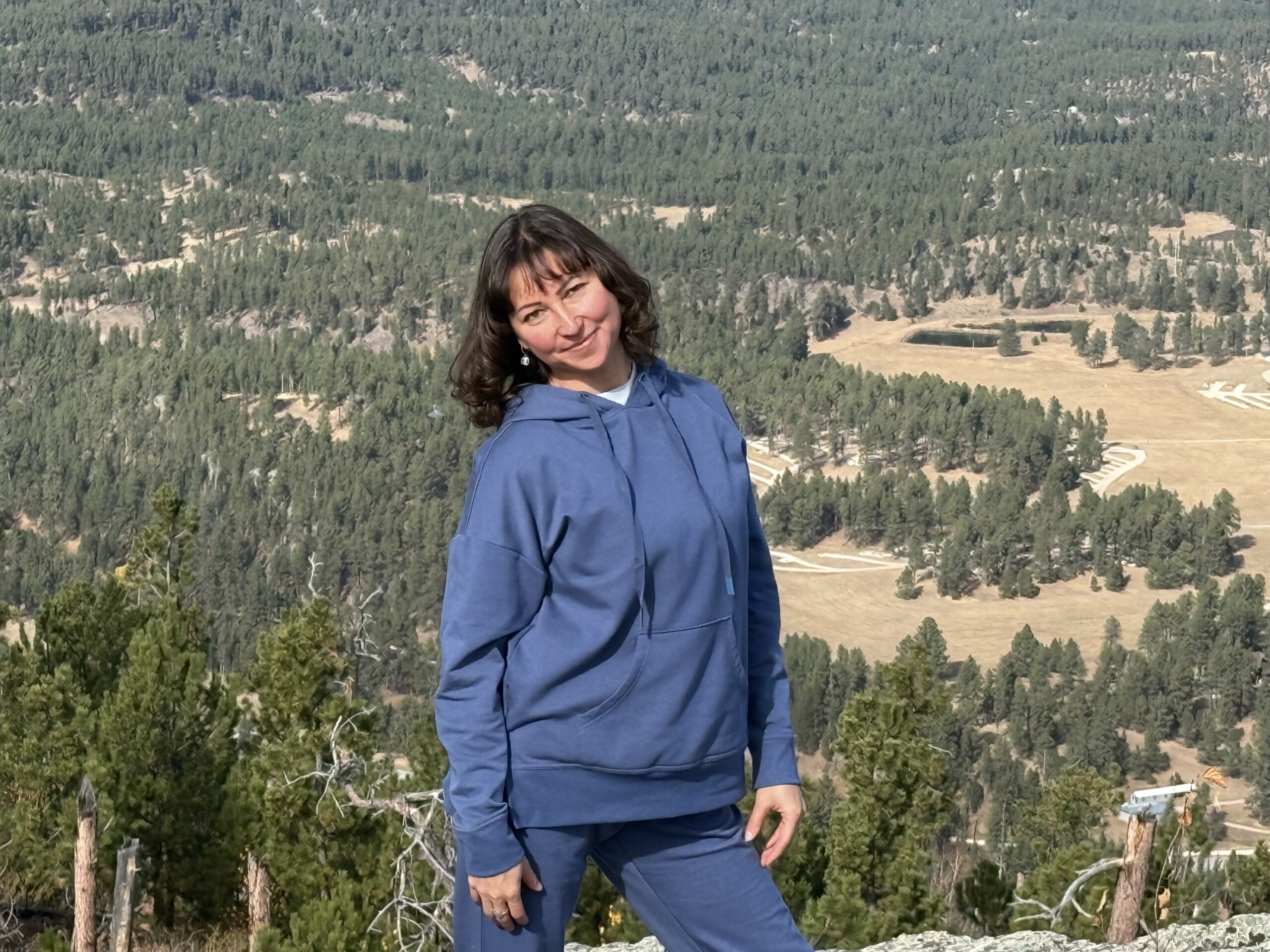 Person in blue hoodie poses in front of a scenic forested mountain landscape.